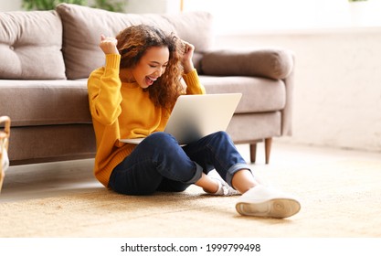 Full length of excited happy afro american woman female student with laptop celebrating success in study, raising arms up and laughing while sitting on floor in living room and studying online at home