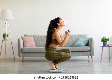 Full length of excited Indian woman sitting on scales at home, overjoyed with success of her slimming diet, side view. Emotional Asian lady achieving her weight loss goal, making YES gesture - Shutterstock ID 1988123912