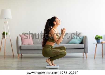 Full length of excited Indian woman sitting on scales at home, overjoyed with success of her slimming diet, side view. Emotional Asian lady achieving her weight loss goal, making YES gesture