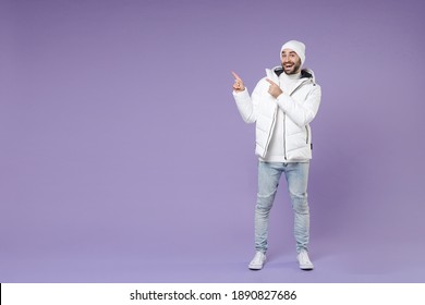 Full Length Of Excited Man In Warm White Padded Windbreaker Jacket Hat Pointing Index Fingers Aside Isolated On Purple Color Background Studio Portrait. People Lifestyle Cold Winter Season Concept