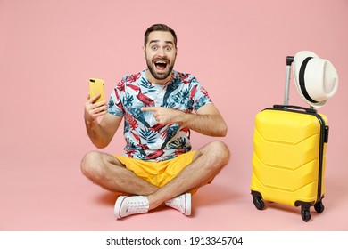 Full Length Excited Traveler Tourist Man Sit On Floor Point Index Finger On Mobile Cell Phone Booking Hotel Taxi Isolated On Pink Background. Passenger Travel On Weekend. Air Flight Journey Concept