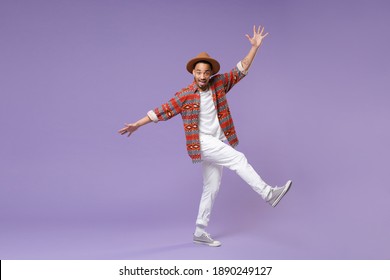 Full Length Excited Young African American Man In Casual Colorful Shirt Hat Dance Rising Leg Spreading Hands Isolated On Violet Background Studio Portrait. People Lifestyle Concept. Mock Up Copy Space