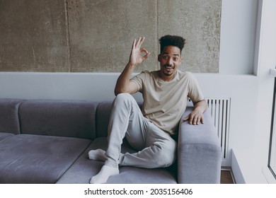 Full length excited young african american man 20s in beige t-shirt sweatpants sitting on comfortbale grey sofa indoors apartment near window show ok okay gesture, resting on weekends staying at home.