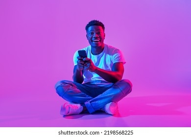 Full Length Of Excited Young African American Guy Sitting Cross Legged With Cellphone, Posting In Social Media, Chatting On Web In Neon Light, Copy Space. Millennial Black Man Using Smartphone