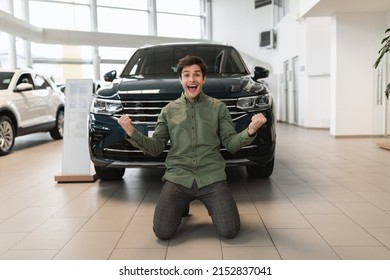Full length of excited young guy shaking fists, making YES gesture, standing on his knees after buying new auto at dealership. Millennial man celebrating purchase of modern vehicle at showroom