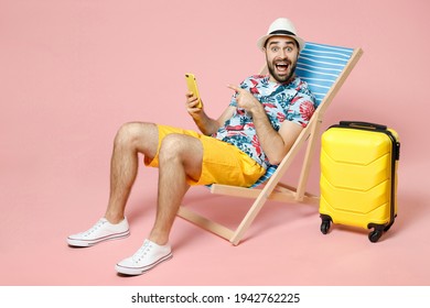 Full Length Excited Young Traveler Tourist Man In Hat Sit On Deck Chair Pointing Index Finger On Mobile Cell Phone Isolated On Pink Background. Passenger Travel On Weekend. Air Flight Journey Concept