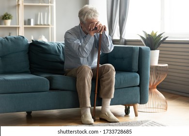 Full Length Exhausted Disabled Middle Aged Old Man Resting On Sofa, Leaning On Walking Cane, Suffering From Depression Alone At Home. Upset Hoary Elder Senior Grandfather Tired Of Hard Rehabilitation.