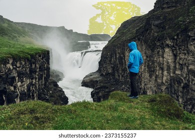 Full length of an explorer standing next to the waterfall and enjoying wild nature. A man is standing on a cliff and looking at the waterfall.