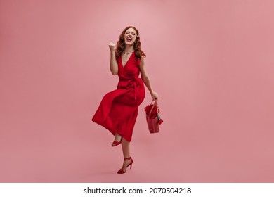 Full Length Fair-skinned Woman Rejoices Over Victory With Open Mouth And Closed Eyes On Pink Background. Red-haired Lady Is Clenching Her Fist, Wearing Red Dress And Shoes Of Same Color.