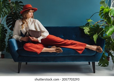 Full length fashion portrait of elegant woman wearing trendy autumn outfit with orange hat, culottes, vintage style blouse, leopard print loafers, holding brown bag, laying on sofa. Copy, empty space
