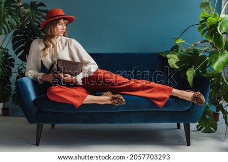 Full length fashion portrait of elegant woman wearing trendy autumn outfit with orange hat, culottes, vintage style blouse, leopard print loafers, holding brown bag, laying on sofa. Copy, empty space
