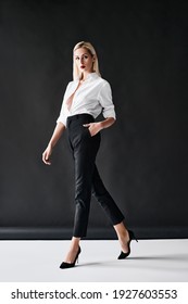 Full length fashion portrait of young stylish woman posing on black background. Trendy blonde female wearing white shirt, black pants and high heel shoes in studio.  