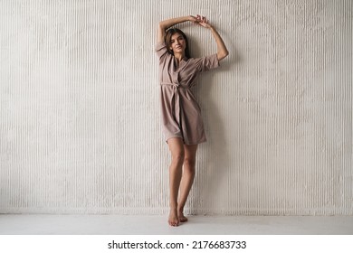 Full Length Fashion Shot Of Adorable Woman In Elegant Silk Dressing Gown, Her Home Outfit, Silk Robe Standing Near Wall In The Morning Advertising New Clothing Collection