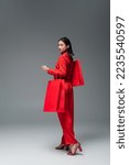 full length of fashionable asian woman with red shopping bags looking away on grey background