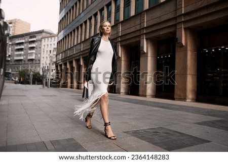 Full length fashionable blonde woman walking city street in chic attire with building in background. Young female model wears white fringed dress, black leather jacket and sandals, looks at camera.