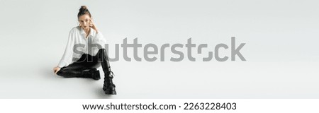 full length of fashionable woman in white shirt and black latex pants posing on grey background, banner