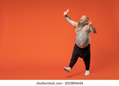 Full Length Fat Fun Pudge Obese Overweight Tattooed Bearded Big Belly Man In Pants With Naked Torso Do Selfie Shot On Mobile Phone Show Victory Gesture Isolated On Orange Background Studio Portrait