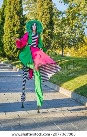 Full length of female clown with painted face in funny costume and green wig, walking on stilts on paved path in park on sunny day