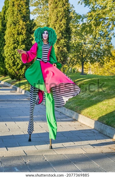 Full length of female clown with painted face in\
funny costume and green wig, walking on stilts on paved path in\
park on sunny day