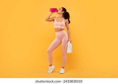 Full length of fit young woman in sportswear drinks water from bottle holding towel, taking workout break over yellow studio background. Healthy hydration during sport training. Free space - Powered by Shutterstock