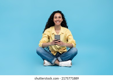 Full Length Fo Cheerful Millennial Lady Sitting Cross Legged With Smartphone, Chatting Online With Friend, Surfing Internet, Watching Video, Posting In Social Media On Blue Studio Background