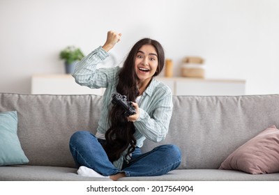 Full length fo excited Indian woman with joystick sitting on couch at home, making victory gesture, winning in video game. Young lady playing online computer arcade, having fun during covid lockdown - Powered by Shutterstock