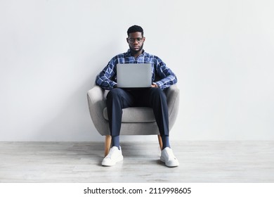 Full Length Of Focused Millennial Black Guy With Laptop Pc Sitting In Armchair, Studying Or Working Online Against White Studio Wall. Serious African American Man Having Remote Business Meeting On Web