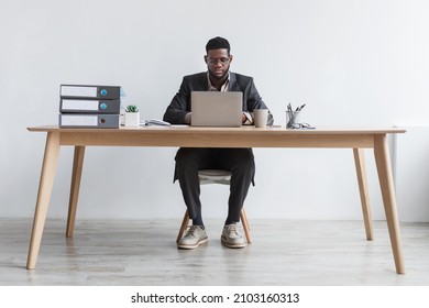 Full length of focused young black man working online with laptop computer, sitting at desk against white studio wall. Serious African American CEO using modern gadget for remote job