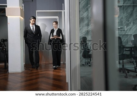 Full length frame of the pair leaving the meeting corner, her eyes on the screen mid stride, his jacket open and stride unhurried in a bright modern coworking office.