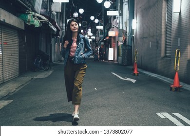 Full Length Front View Of Asian Woman With Backpack Walking On Quiet Street At Night With No City Traffic Car Trails. Female Back To Home Alone In Alley. Beautiful Lady Hand In Pocket Smiling Relax.