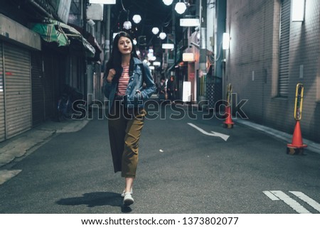 full length front view of asian woman with backpack walking on quiet street at night with no city traffic car trails. female back to home alone in alley. beautiful lady hand in pocket smiling relax.