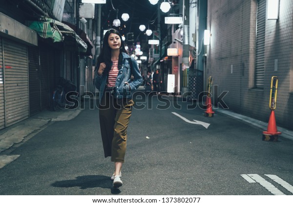 full length front view of asian woman with\
backpack walking on quiet street at night with no city traffic car\
trails. female back to home alone in alley. beautiful lady hand in\
pocket smiling relax.