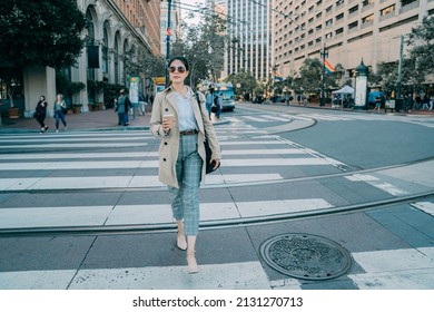 Full Length Front View Beautiful Asian Korean Woman Crossing On Zebra Crosswalk And Holding Take Away Coffee In Hand. Smiling Young Office Lady Relax Walking In City Street. Gay Flag Hanging Usa.