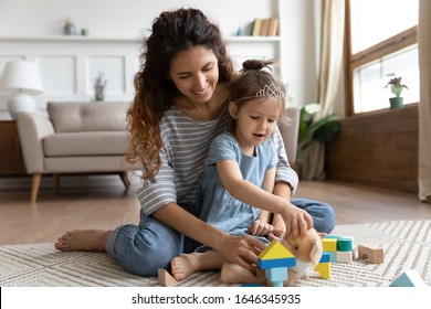 Full length front view happy young mother cuddling small preschool kid daughter, playing with fluffy toys and colored wooden bricks on floor carpet in living room. Smiling nanny having fun with child. - Shutterstock ID 1646345935
