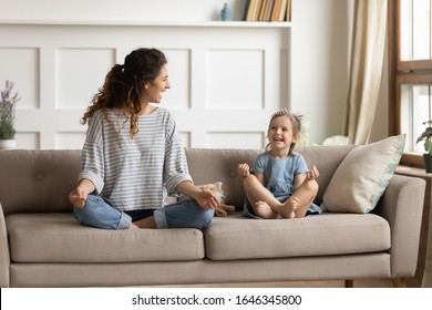 Full length front view smiling young curly mother sitting on comfortable couch with cute playful little preschool daughter in lotus position. Happy mommy practicing yoga exercised with small child. - Powered by Shutterstock