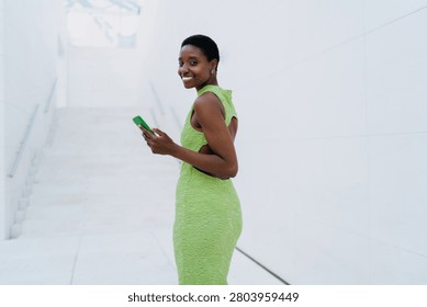 Full length front view of a stylish African American woman in a green maxi dress and white sneakers, leaning on a modern metal mesh fence and text messaging on a smartphone. She stands against a clean white architectural wall under bright daylight.