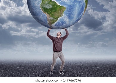 Full length front view of young man in casual clothes holding up big Earth globe on gloomy overcast day. Use of natural resources. Environmental issues. Trying to help planet.
