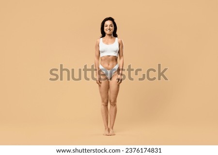 Full length frontal view of slim woman with perfect body shape and flat tummy posing on beige studio background, attractive lady wearing white underwear looking and smiling at camera, copy space