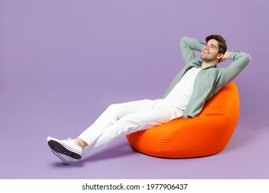 Full length fun man 20s in casual mint shirt white t-shirt sitting in orange bean bag chair hold hands folded behind neck isolated on purple color background studio portrait People lifestyle concept - Powered by Shutterstock