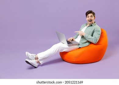 Full Length Fun Man 20s In Casual Mint Shirt White T-shirt Sitting In Orange Bean Bag Chair Point Finger On Laptop Pc Computer Chat Isolated On Purple Color Background Studio People Lifestyle Concept