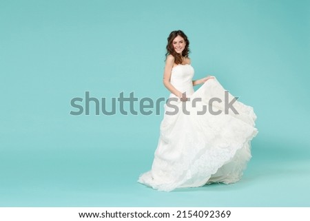Full length fun smiling tender bride woman 20s in beautiful white wedding dress standing looking camera isolated on blue turquoise color background studio portrait. Ceremony celebration party concept