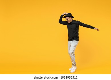 Full length fun young american african man 20s wears stylish black hat shirt eyeglasses touching hat dancing have fun with outstretched hand isolated on yellow orange color background studio portrait.