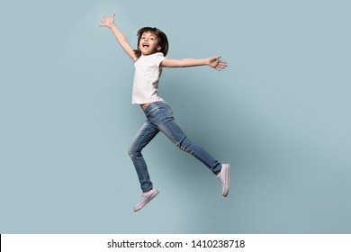 Full length funny active six years old little girl wearing casual clothes jeans white t-shirt gumshoes flying jumping with raised hands happy positive mood emotions isolated on blue studio background - Shutterstock ID 1410238718