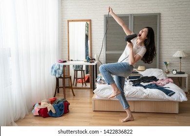 full length of funny asian girl dancing and singing with hair dryer in wood cozy bedroom at home. young chinese woman crazy joyful with clothes dresses on bed prepared for luggage on sunshine day.