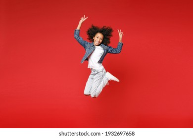 Full Length Of Funny Little African American Kid Girl 12-13 Years Old In Denim Jacket Jumping Showing Victory Sign Isolated On Red Background Children Studio Portrait. Childhood Lifestyle Concept