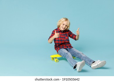 Full length of funny little curly kid boy 10s in basic red checkered shirt sitting on skateboard showing thumbs up isolated on blue background children studio portrait. Childhood lifestyle concept