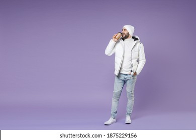 Full Length Funny Man In Warm White Windbreaker Jacket Hat Hold Paper Cup Coffee Tea Drinking Looking Aside Isolated On Purple Background Studio Portrait. People Lifestyle Cold Winter Season Concept