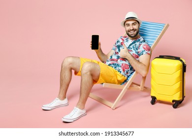 Full Length Funny Tourist Man In Hat Sit On Deck Chair Hold Mobile Phone With Blank Empty Screen Showing Thumb Up Isolated On Pink Background. Passenger Travel On Weekend. Air Flight Journey Concept