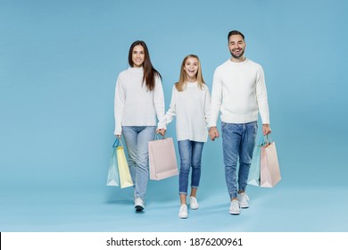 Full length of funny young parents mom dad with child kid daughter teen girl in sweaters hold package bags with purchases after shopping isolated on blue background. Family day parenthood concept