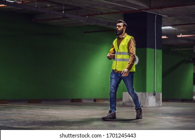 Full Length Of Good-looking Bearded Worker In Vest, With Safety Helmet Under Armpit Walking Around Underground Parking Lot In Construction Process.